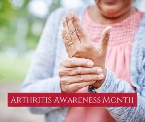 A person is gently massaging their hand, possibly indicating joint pain. They are wearing a pink top and gray cardigan. Text on the image reads Arthritis Awareness Month. The background is a blurred outdoor setting.